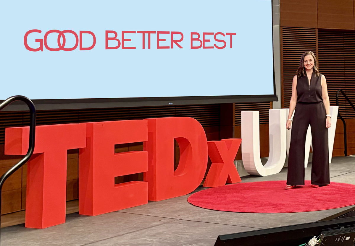 Angela, the founder, stands on a red circular rug on a TEDx stage, with large red and white TEDx letters and a screen behind her reading 'GOOD BETTER BEST'.