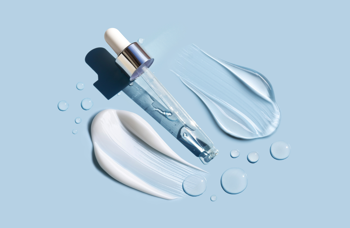 A clear glass skincare dropper bottle with a white dropper and silver collar, arranged on a light blue background with white and translucent cream swatches and water droplets.
